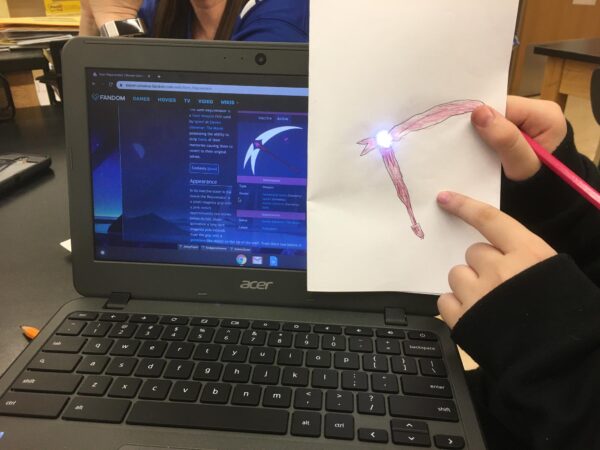 A student’s hand holds a folded piece of paper with a hand drawn light up sword in front of a laptop. The laptop shows a digital illustration of this same sword.