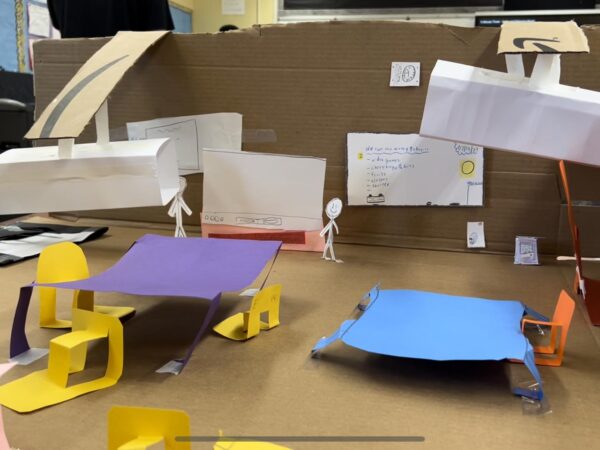 A miniature replica of a classroom. The walls are made out of cardboard, the tables are made out of a purple and a blue piece of paper. There are yellow and orange paper chairs, two paper projectors, three white boards, and two paper people at the front of the room.