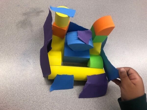 A tower with a square base made out of yellow, blue, orange and green foam blocks. Ripped blue and purple pieces of paper are attached along the sides.