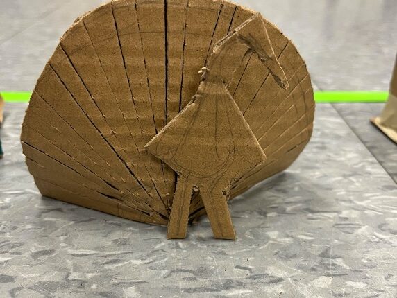 A mini cardboard peacock. The peacock’s feathers are on display and is made from a semi-circle shaped piece of scored cardboard. It rests behind a cardboard outline of the peacock’s body in profile.