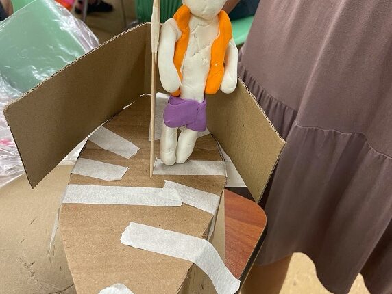 A cardboard boat held together with masking tape. A narrow wooden dowel is in the center and a clay person wearing an orange vest and purple shorts stands beside it.