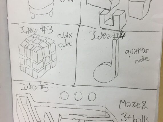 The inside of a student’s white paper journal is shown where there are multiple ideas for different puzzles and shapes to make. Some ideas are of a Rubik's Cube, a quarter note and a maze with three balls.
