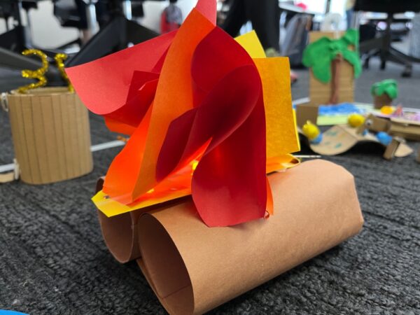A paper bonfire made out of rolled brown paper for the base and red, yellow, and orange paper crinkled paper at the top. An illuminated LED is tucked in between the colored paper.