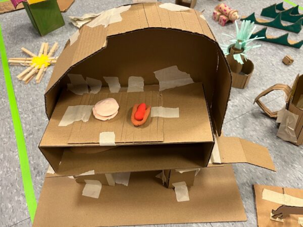 A mini cardboard grill made from scored pieces cardboard, taped together with masking tape. The grill is exposed and shows a hot dog made from red playdough and a burger made from cream-colored playdough.