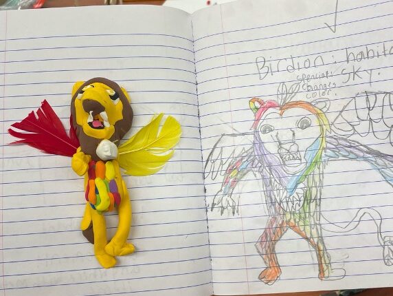 A yellow "birdion" figure made out of yellow and brown clay. There are multi-colored clay feathers at the torso and a red and a yellow feather for wings tucked behind the figure. It sits on the left side of an open journal page with a student’s sketch of the figure on the right.