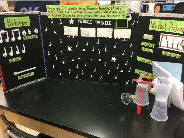 A student’s final project is presented on a black poster board with neon green paper labels. On the left side of the board, there is a 3D printed score of Twinkle, Twinkle Little Star with prototype versions and the perfected 3D musical notes. On the right side there are samples of other completed projects such as a 3D eraser, a circuit board, journal and a vibrating motor.