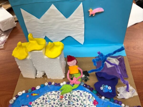 A student’s final project of a mermaid world. A mermaid made of playdough and pipe cleaners sits by a body of water made from beads, clay paper, and stickers.The cardboard base includes a styrofoam castle. The cardboard background is covered in a light blue sheet of paper and a white piece of paper cut out to look like waves is mounted on top. A small sea creature is glued beside the waves.