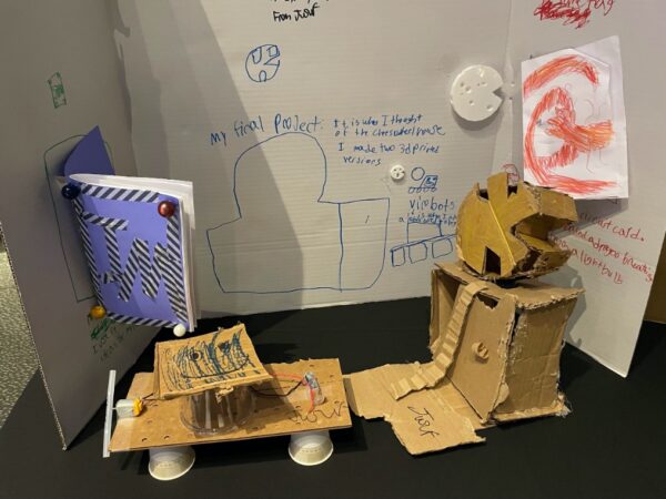 Final project showing several artifacts, including a handmade journal, a light-up card with a dragon whose fire-breathing capacity is fueled by an LED, a cardboard construction of a cheese wheel house, and a drawn and 3D printed versions of the cheese wheel itself. In the foreground is a recycled-materials vibrabot, a bot whose movement comes from the shaking generated by an off-center weight (the hot glue stick) as it spins with the rotating motor shaft. Sewing, cardboard cutting, circuit making, and 3D printing