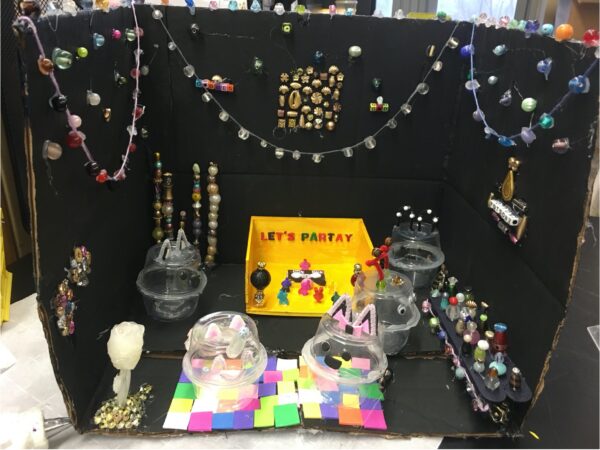 Strands of beads are festooned across the top halves of each section of a black cardboard trifold. The bottom has a yellow dance floor with 3D printed characters and letters that spell Let’s Party. Cat faces made from clear plastic cuts sit on a foam squares dance floor. A long table with bottles made from beads serve as the snack area.
