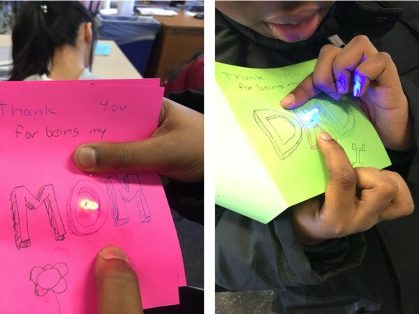 The picture on the left shows a student hand holding a paper circuit. There is a rectangle of copper tape with a strip of tape across the middle. In the middle of the top and center lines are single lit LEDs. The picture on the right shows a red card. The card reads Mom. The picture on the left shows a green card. The card reads Dad.