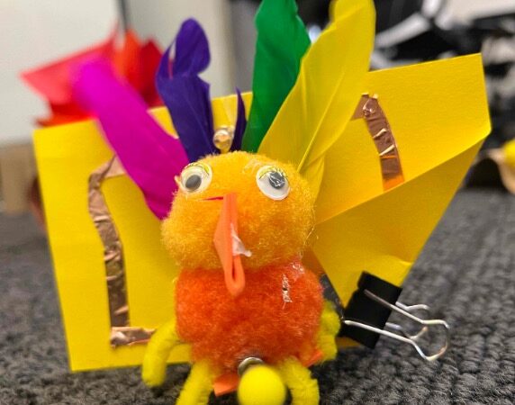 A small bird made of a yellow and an orange pompom, colorful feathers, googly eyes, and a felt beak. A yellow piece of paper with copper tape sits behind it.